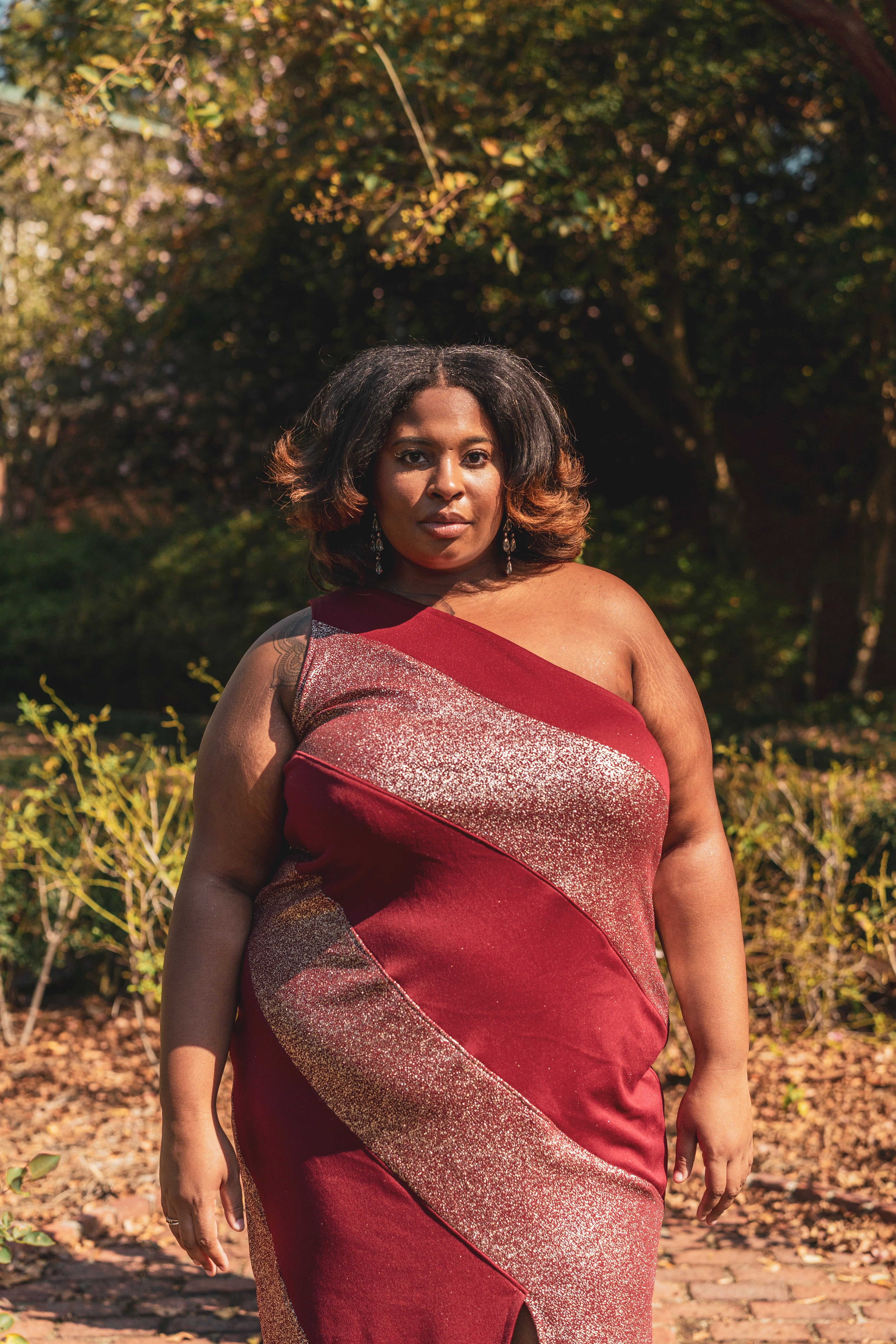 Woman wearing a red and gold dress standing outdoors with greenery in the background