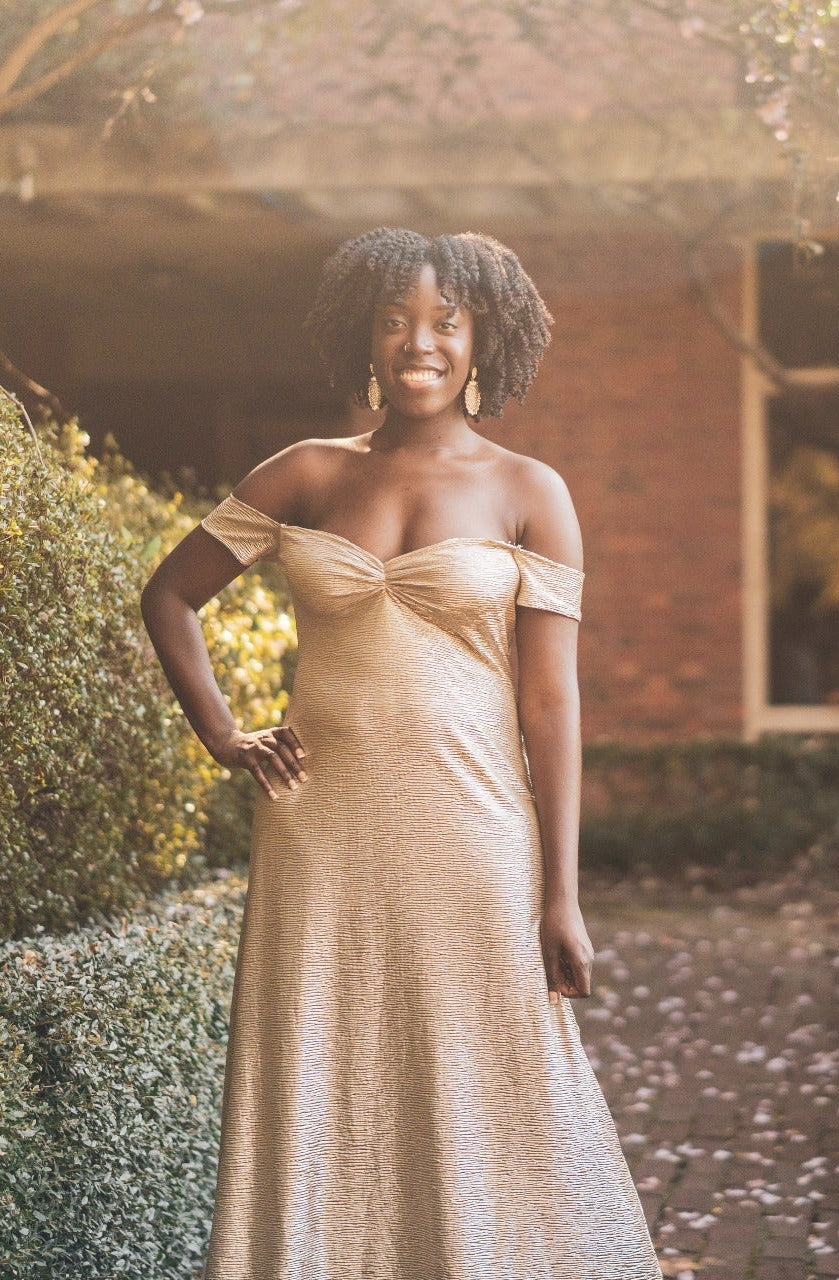 Woman in a metallic biege off-the-shoulder dress standing in a garden with a brick building in the background