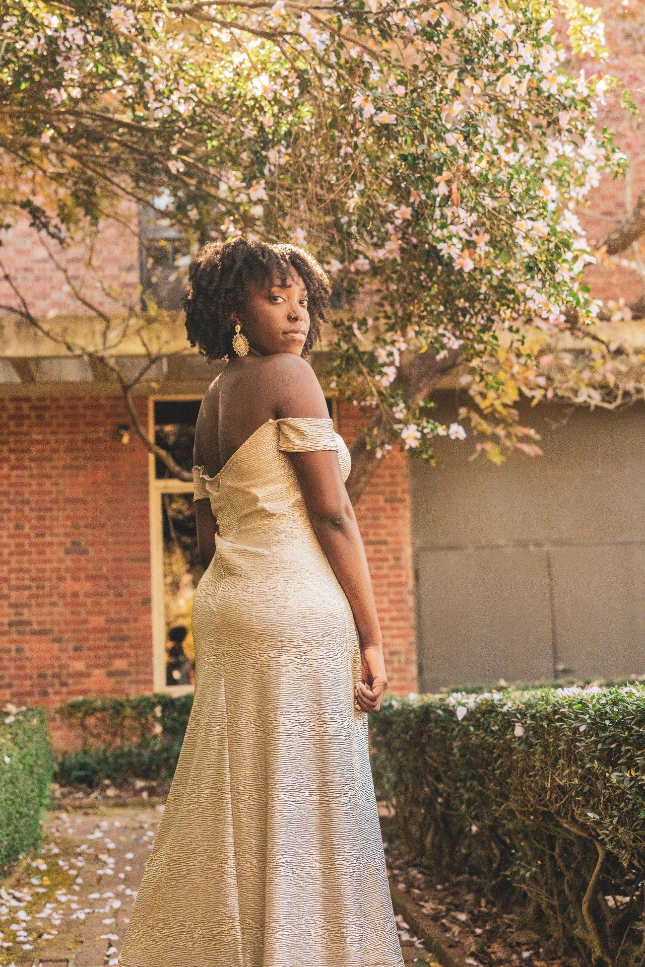 Woman in a metallic beige off-shoulder dress standing under a tree with blossoms.